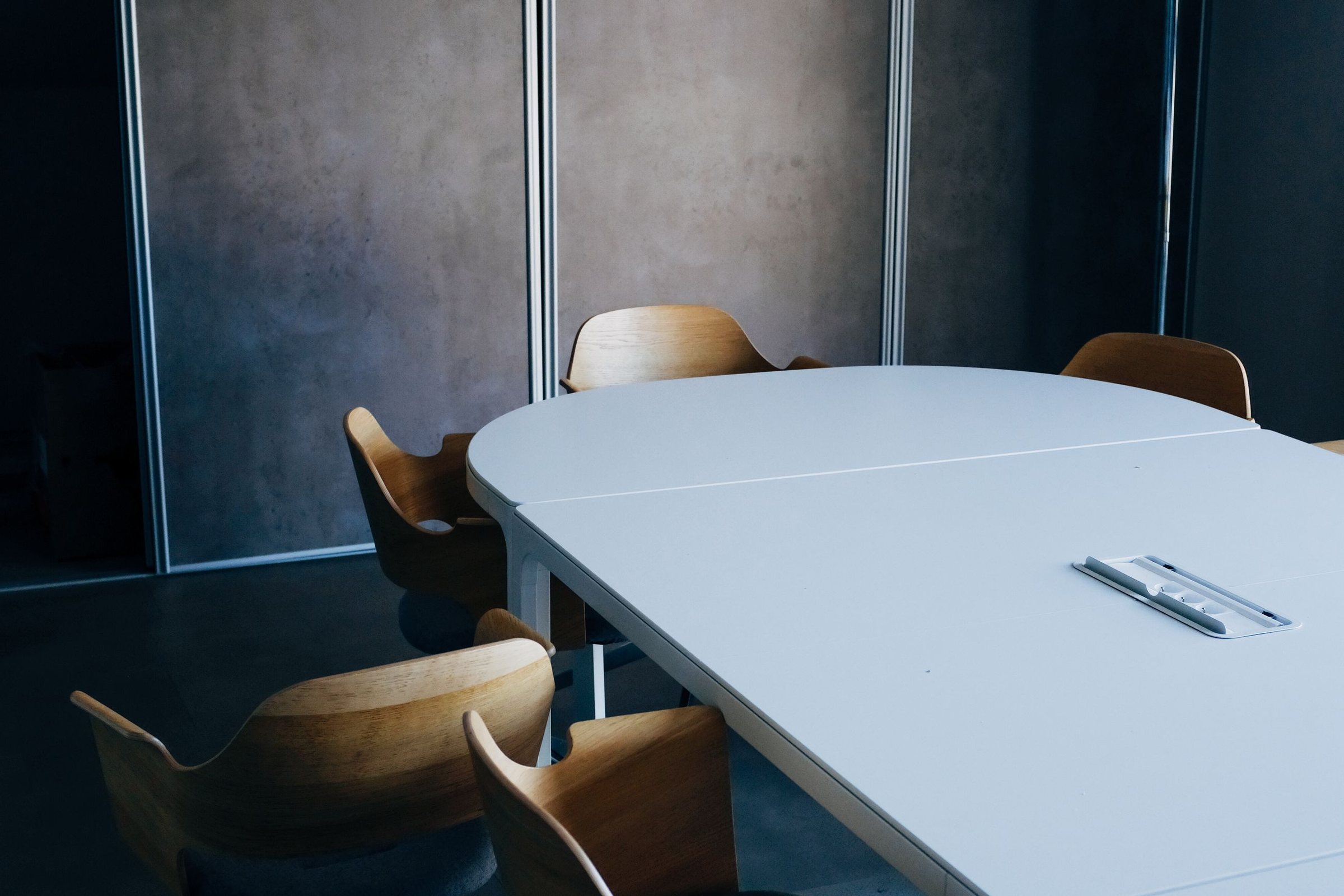 An empty meeting room with a pale oval table, bentwood chairs and plain plaster wall panels in daylight.