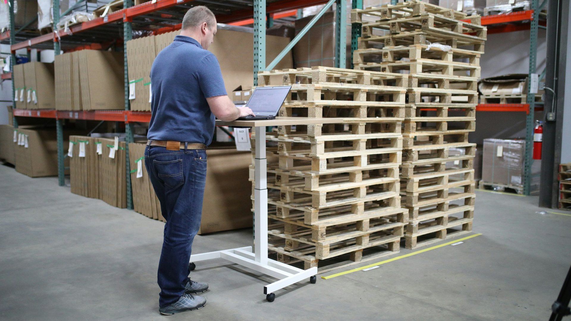 A man in a blue polo shirt working at a laptop on a height-adjustable standing desk beside stacked wooden pallets in a racked warehouse