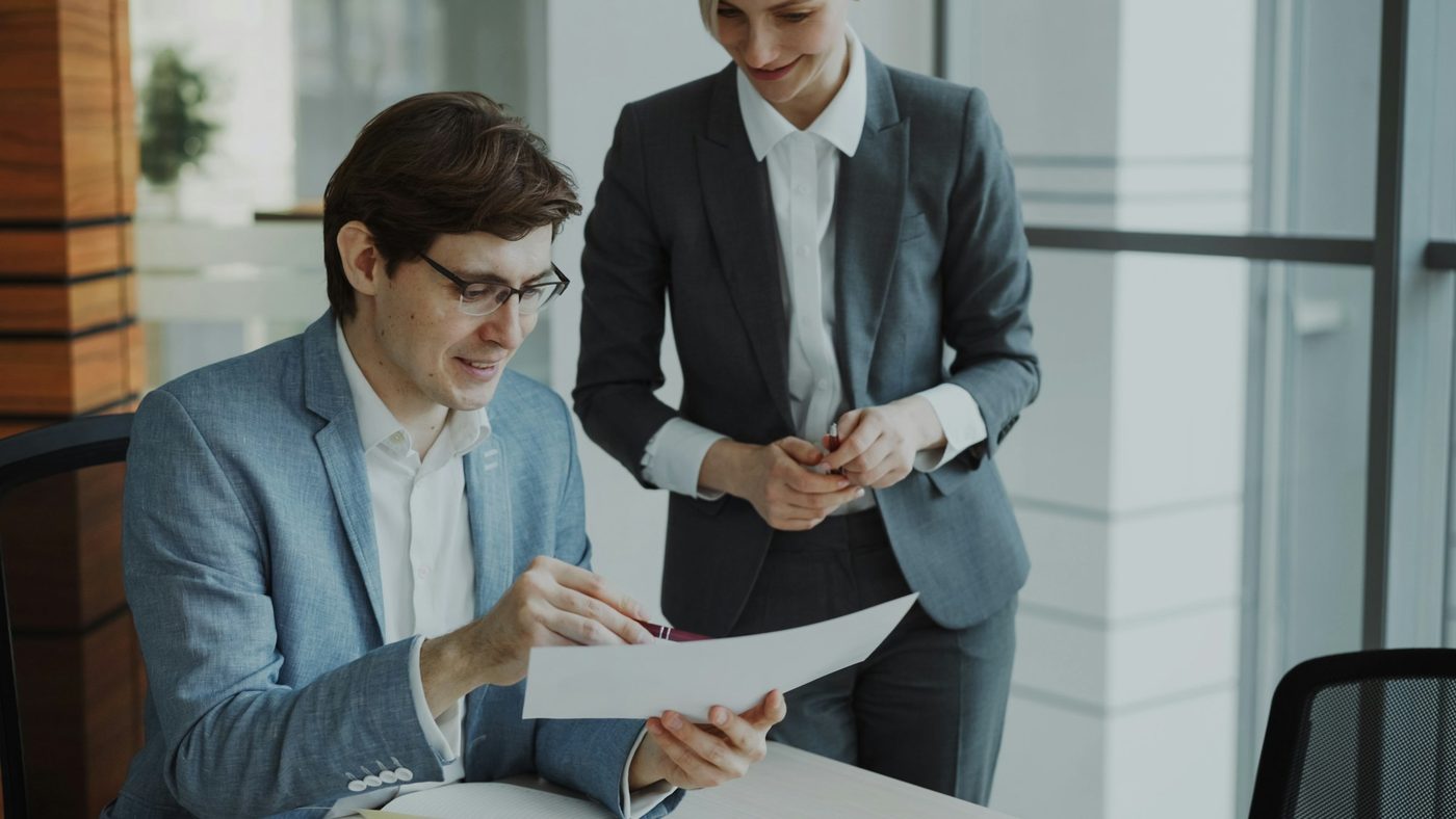 Illustrative stock photo: a man seated at a desk by a window points with a pen at a printed report while a colleague stands beside him and listens.