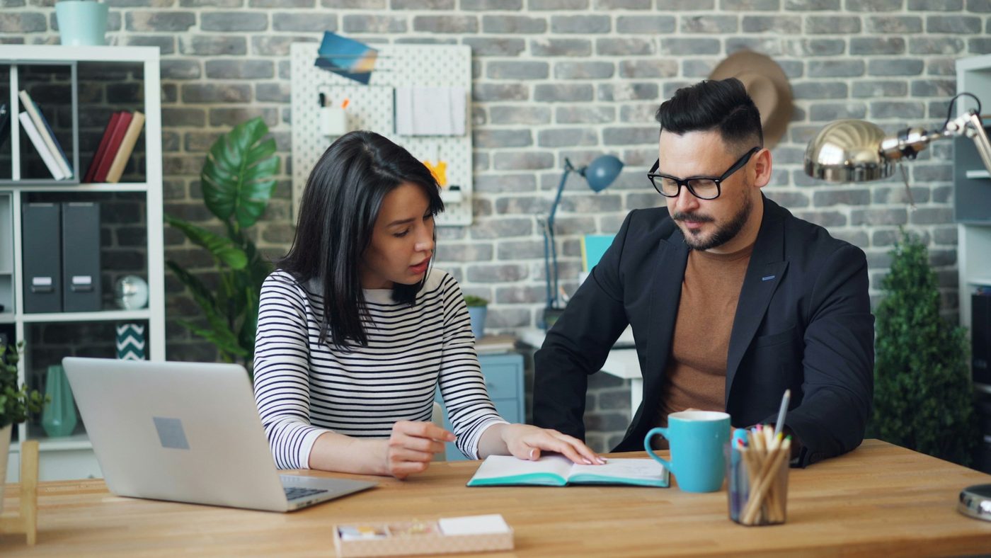 Two colleagues at a shared office desk. One points to a line in an open notebook while the other reads along.