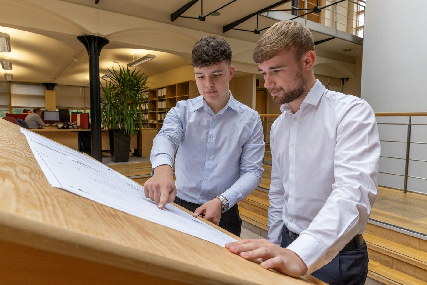 Illustrative stock photograph: two colleagues in a UK office leaning over a large printed technical drawing on a wooden desk, one pointing at a detail on the sheet.
