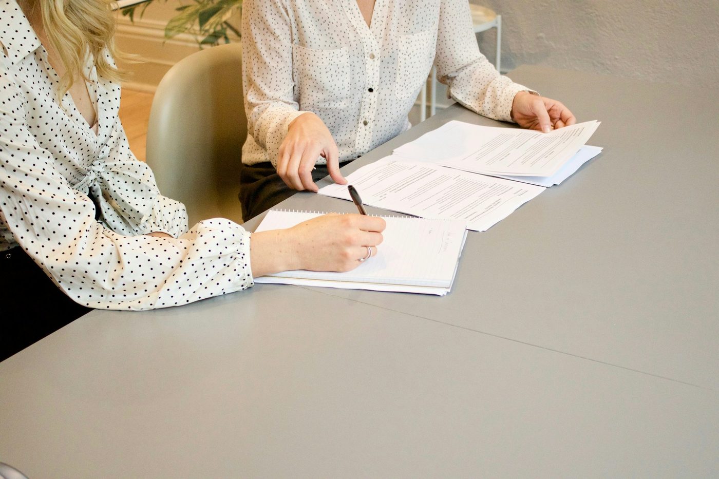 Illustrative stock photo: two people at a plain grey meeting table go through printed pages; one points at the document while the other writes in a notebook, with faces out of frame.