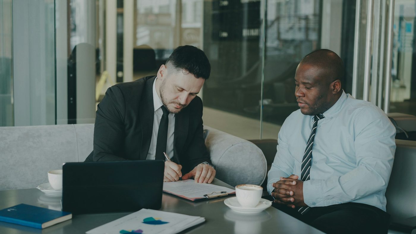 Two colleagues sit side by side at a small table. One writes on a printed document on a clipboard while the other leans in to read it.
