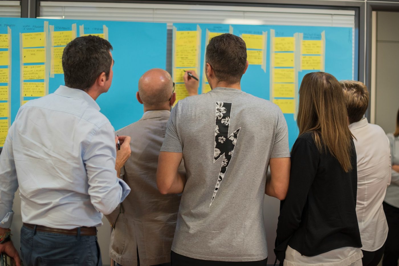 A group of colleagues, seen from behind, gathers at a wall of paper marked out in lanes of handwritten sticky notes while one of them adds a note.