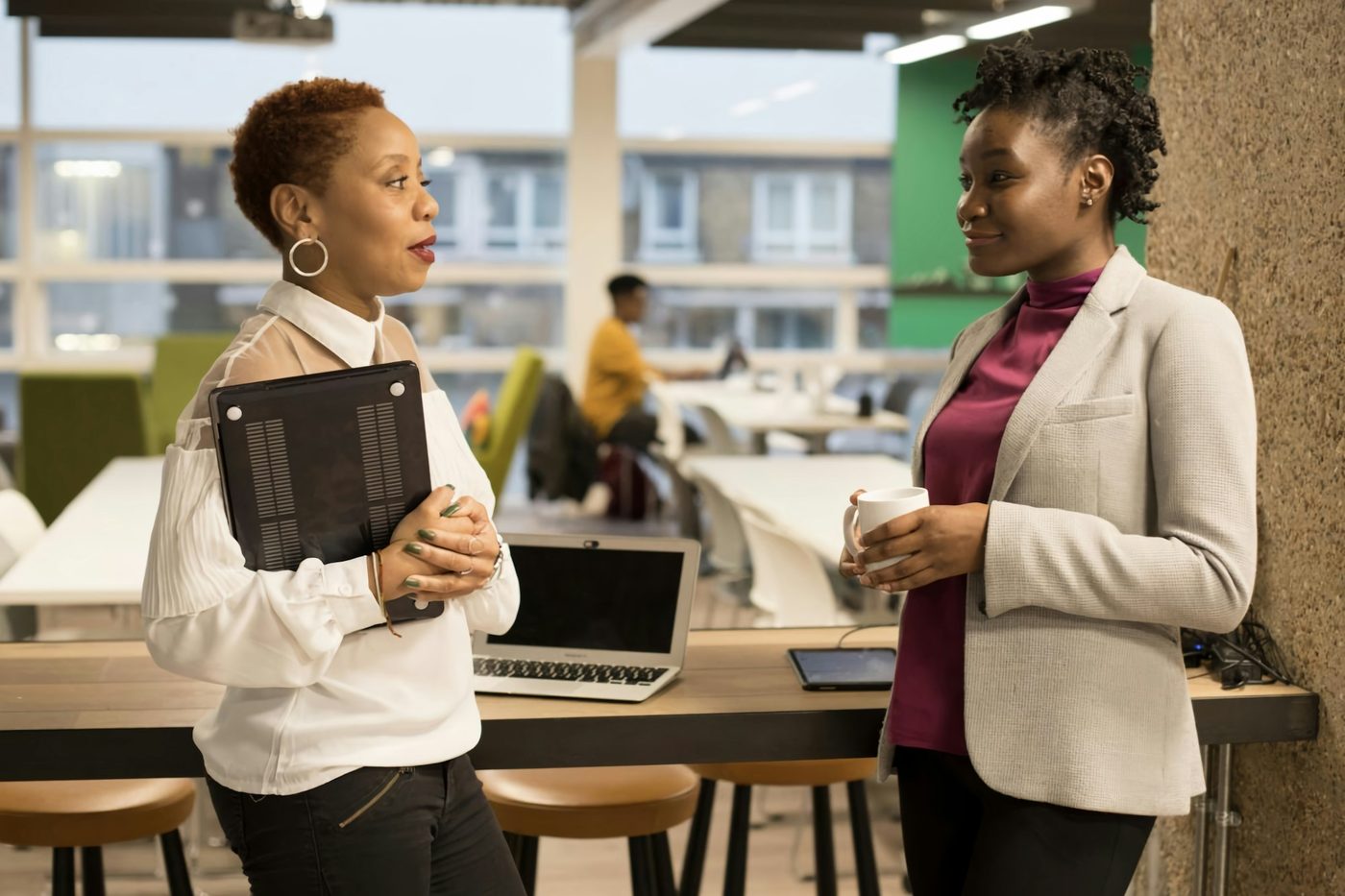 Illustrative stock photo: two colleagues talk standing by the windows of an open-plan office. One holds a closed laptop against her chest like a folder, the other holds a mug. Ordinary desks and a colleague at work are behind them.