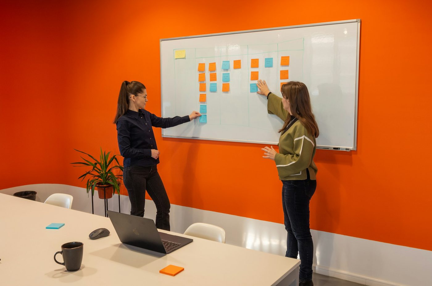 Illustrative stock photo: two colleagues in a small meeting room move sticky notes between hand-drawn columns on a whiteboard. A mug and a laptop sit on the table in the foreground.