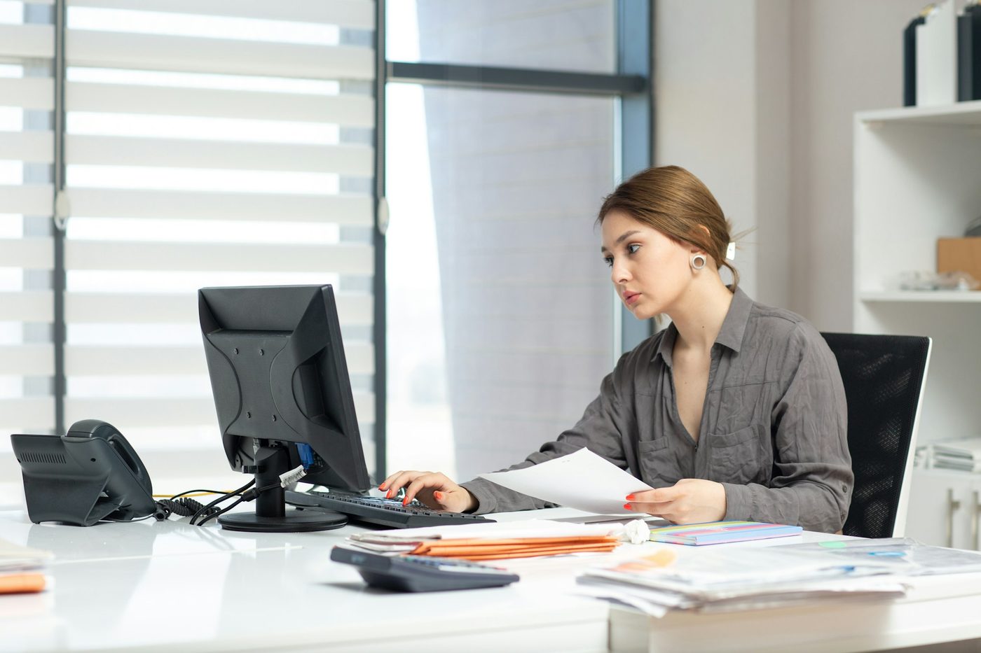 Illustrative stock photo: an office administrator types into a desktop computer while reading from a printed sheet, with piles of paper records, folders and a calculator on the desk. The monitor faces away from the camera.