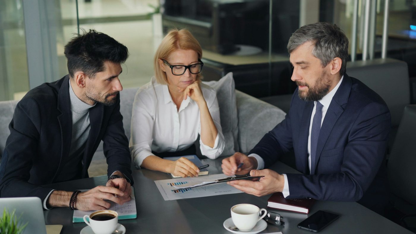 Illustrative stock photo: three colleagues at a small table lean in while one points with a pen at a printed bar chart, with coffee cups and notebooks around them.