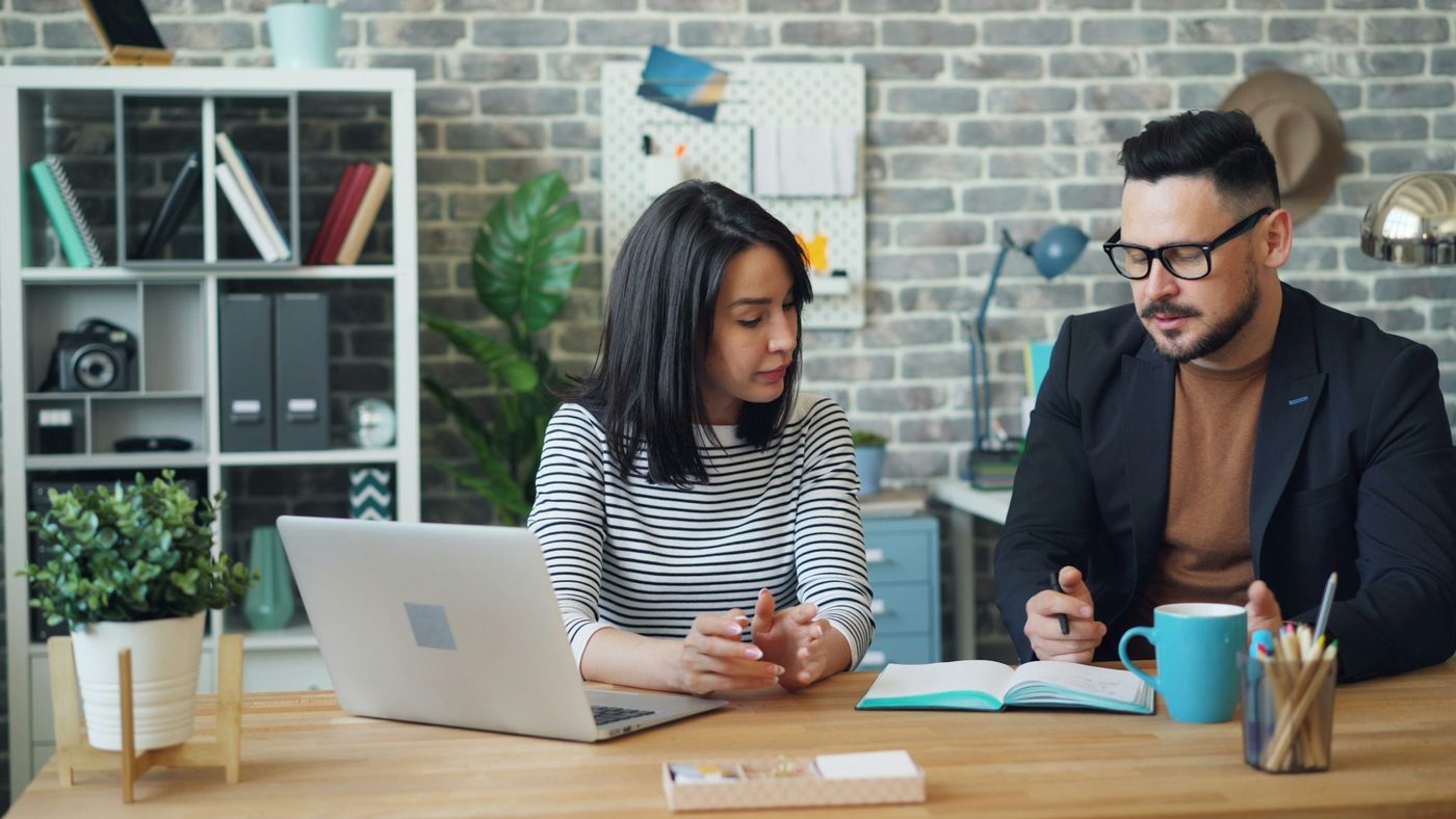 Illustrative stock photo: two colleagues at a shared wooden desk. One explains something beside an open laptop angled away from the camera, while the other takes notes in a paper notebook.