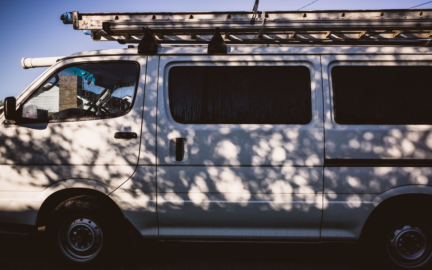 A plain white works van seen side on in late afternoon sun, with a paint-spattered ladder strapped to its roof rack and tree shadow across the panels.