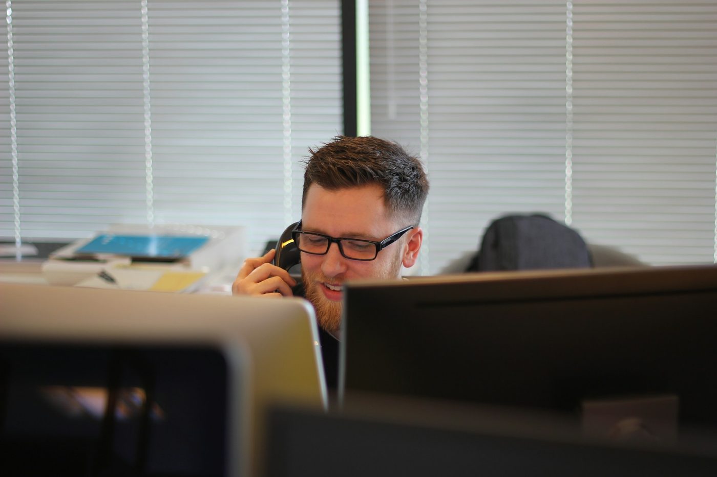 Illustrative stock photo: a man in glasses takes a call on a desk phone behind a row of monitors turned away from the camera, with window blinds behind him in an ordinary office.