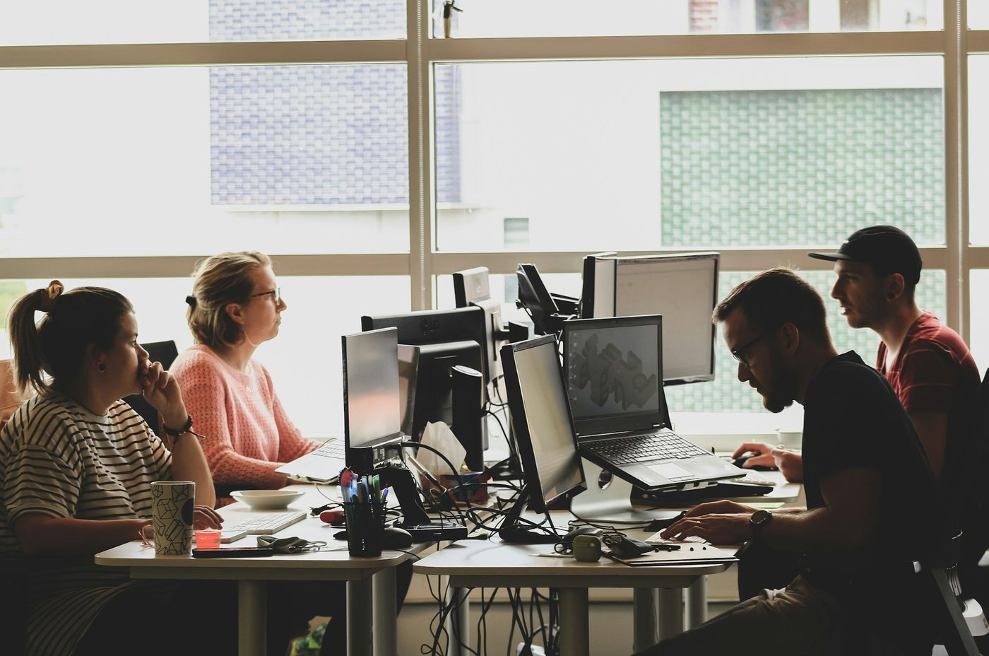 Illustrative stock photo: four colleagues work at adjoining desks by a large window in a small office, two of them talking across the monitors, with mugs and pens on the desks.