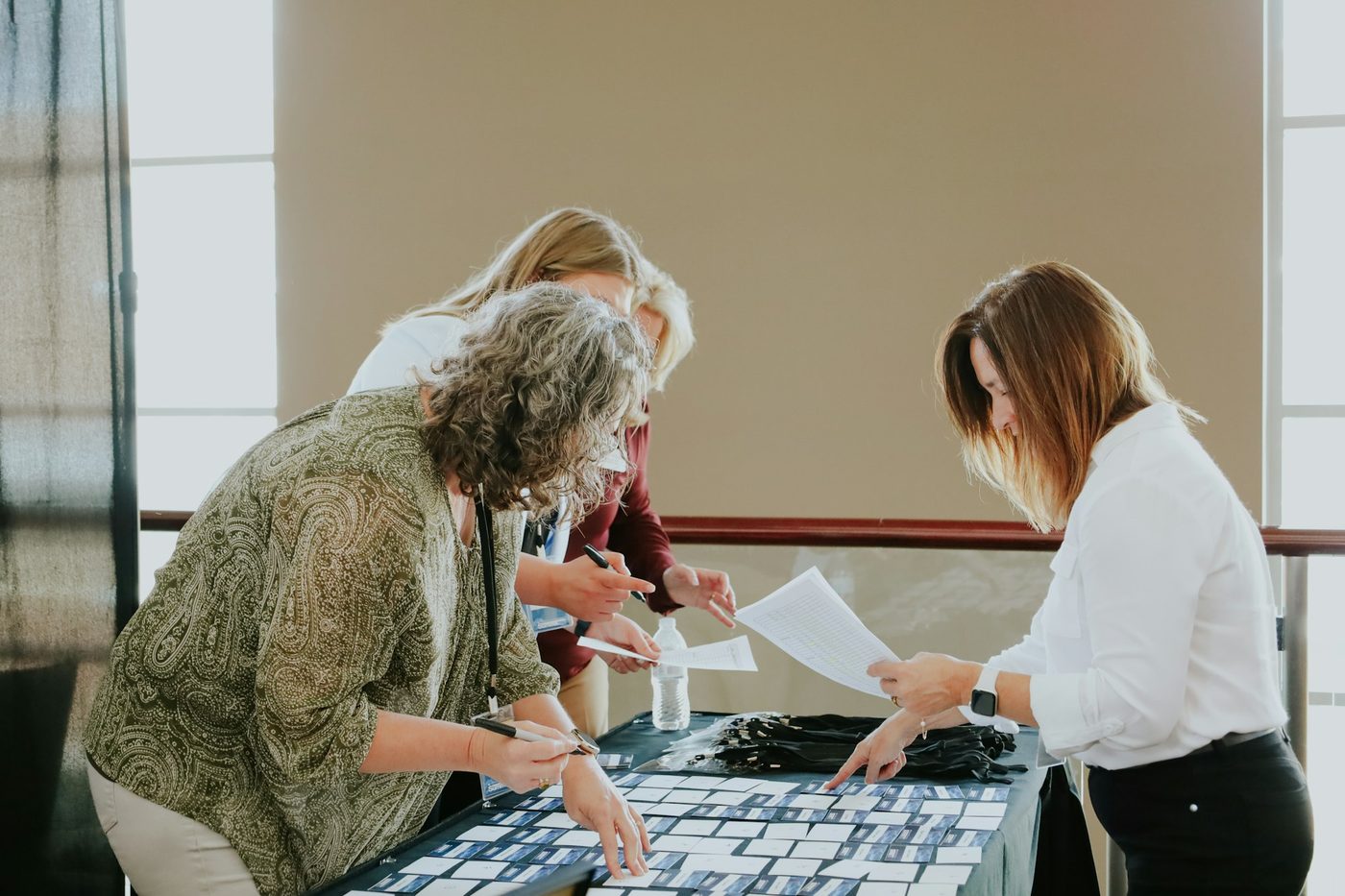 Three colleagues lean over a table in window light, sorting rows of printed cards and checking printed lists by hand; one wears a lanyard.
