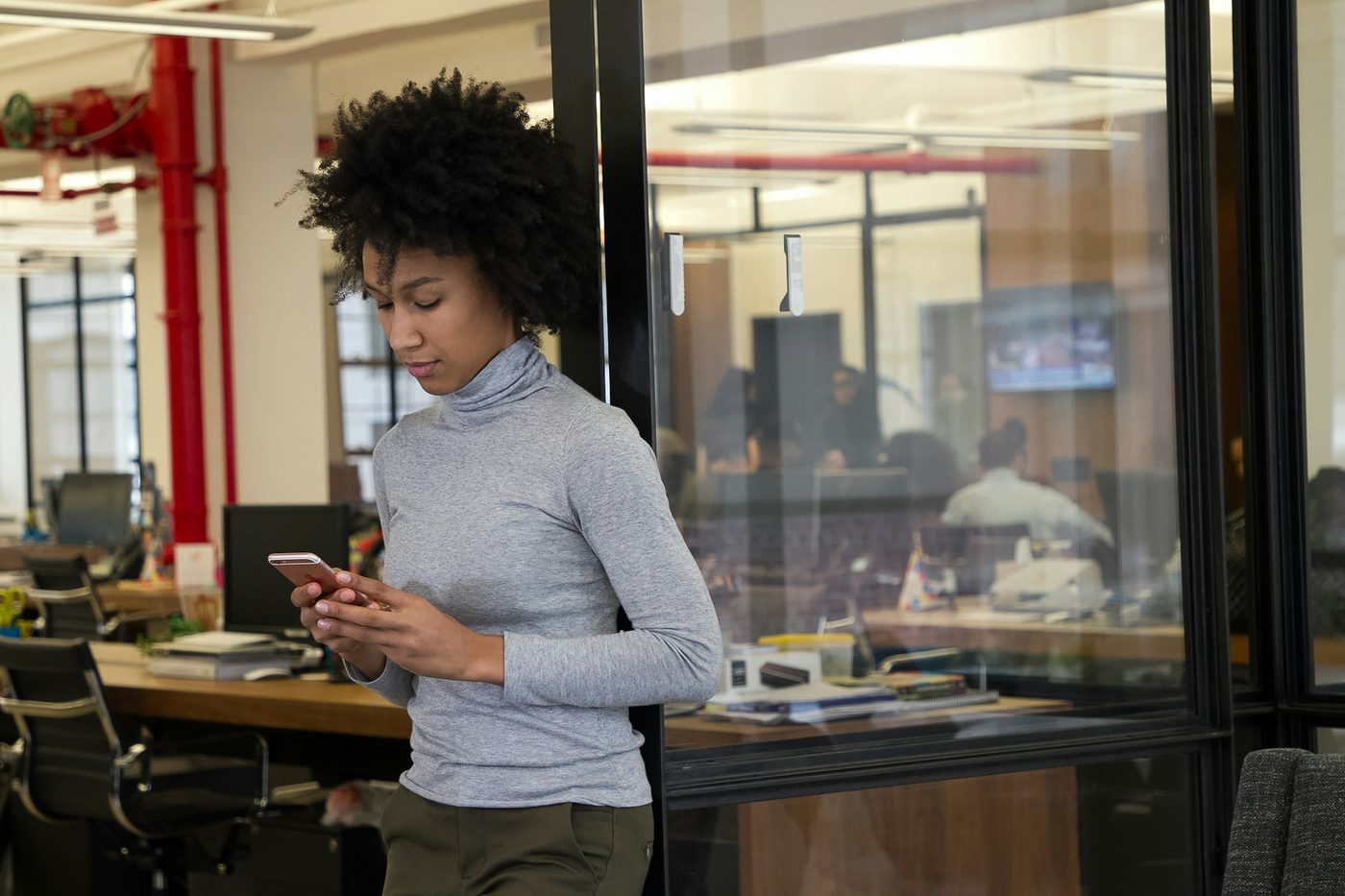 Illustrative stock photo: a woman stands by a glass partition in an open-plan office, reading something on her phone, with desks and a meeting room behind her.
