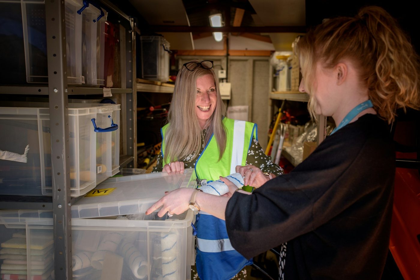 Illustrative stock photo: in a cramped UK stockroom, a woman in a hi-vis vest and an office colleague wearing a lanyard check paint rollers from a plastic storage crate between shelving units.