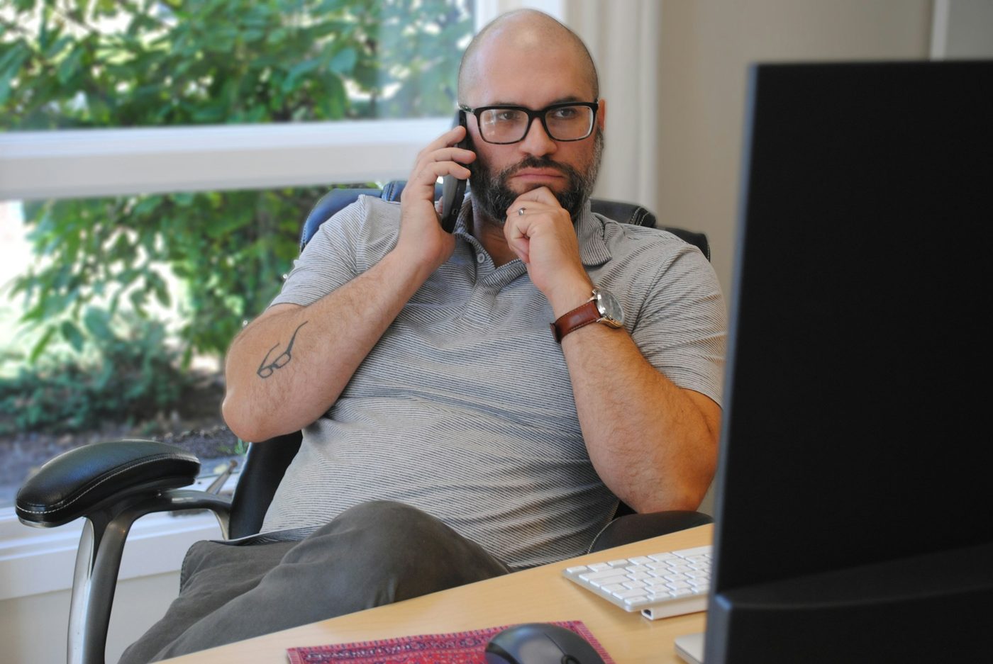 A man at his desk takes a phone call in daylight, with a window onto greenery behind him and his monitor turned away from the camera.