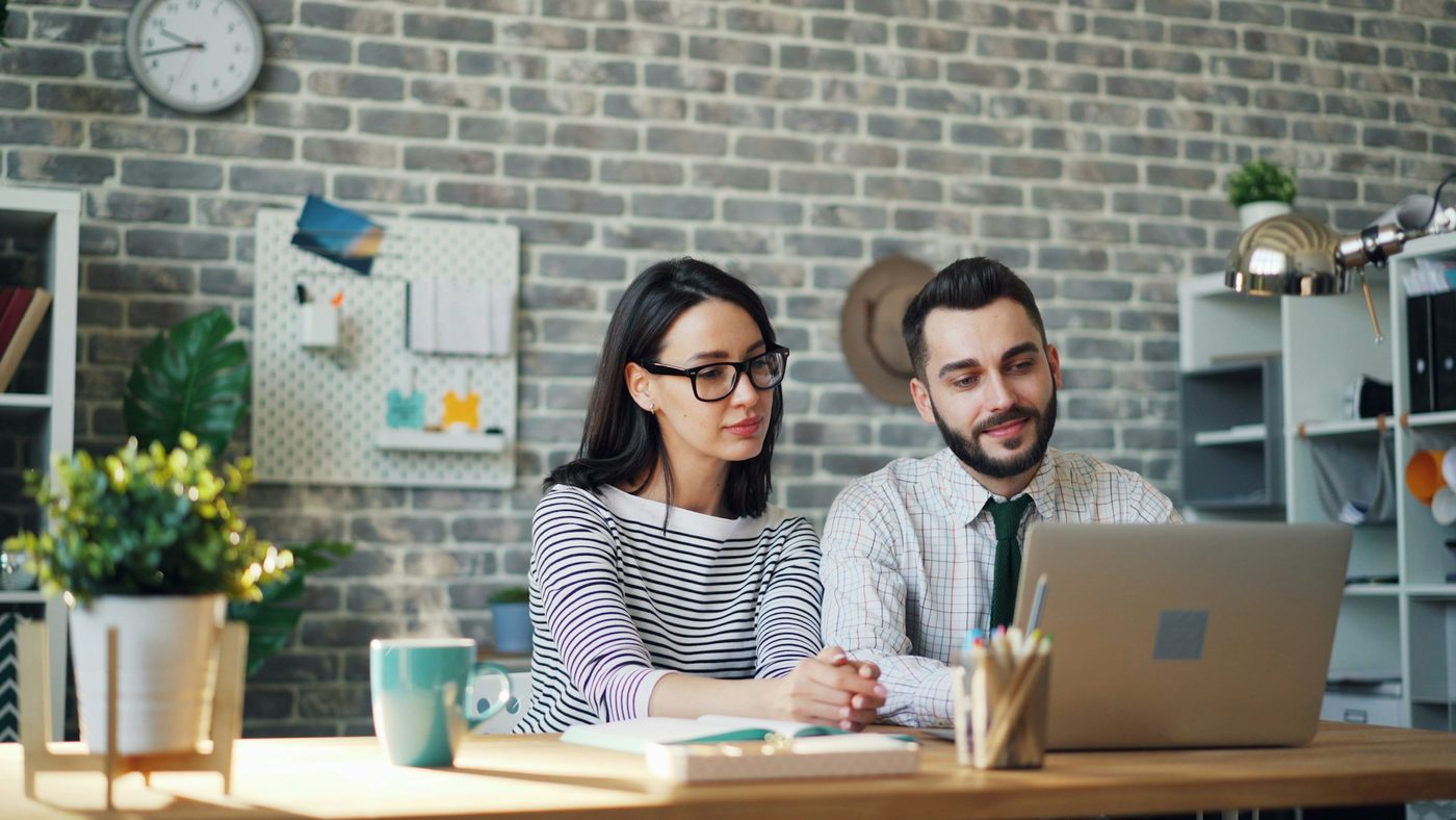 Illustrative stock photo: two colleagues sit side by side at a desk in a small office with a grey brick wall. They look at a laptop whose screen faces away from the camera, with a mug and a notebook on the desk.
