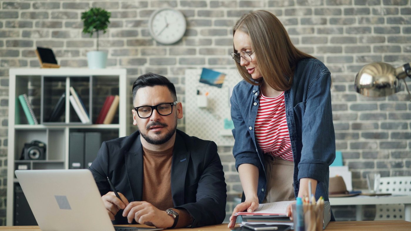 A seated colleague at a laptop and a standing colleague holding an open ring binder, going through a printed page together in a brick-walled office