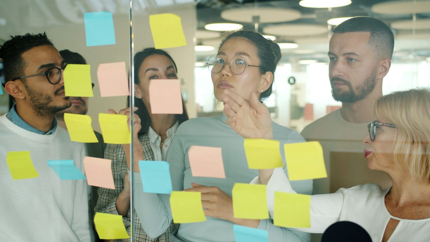 Illustrative stock photo: a small team stands behind a glass wall covered in sticky notes arranged in loose columns. One colleague gestures at a note while the others watch.