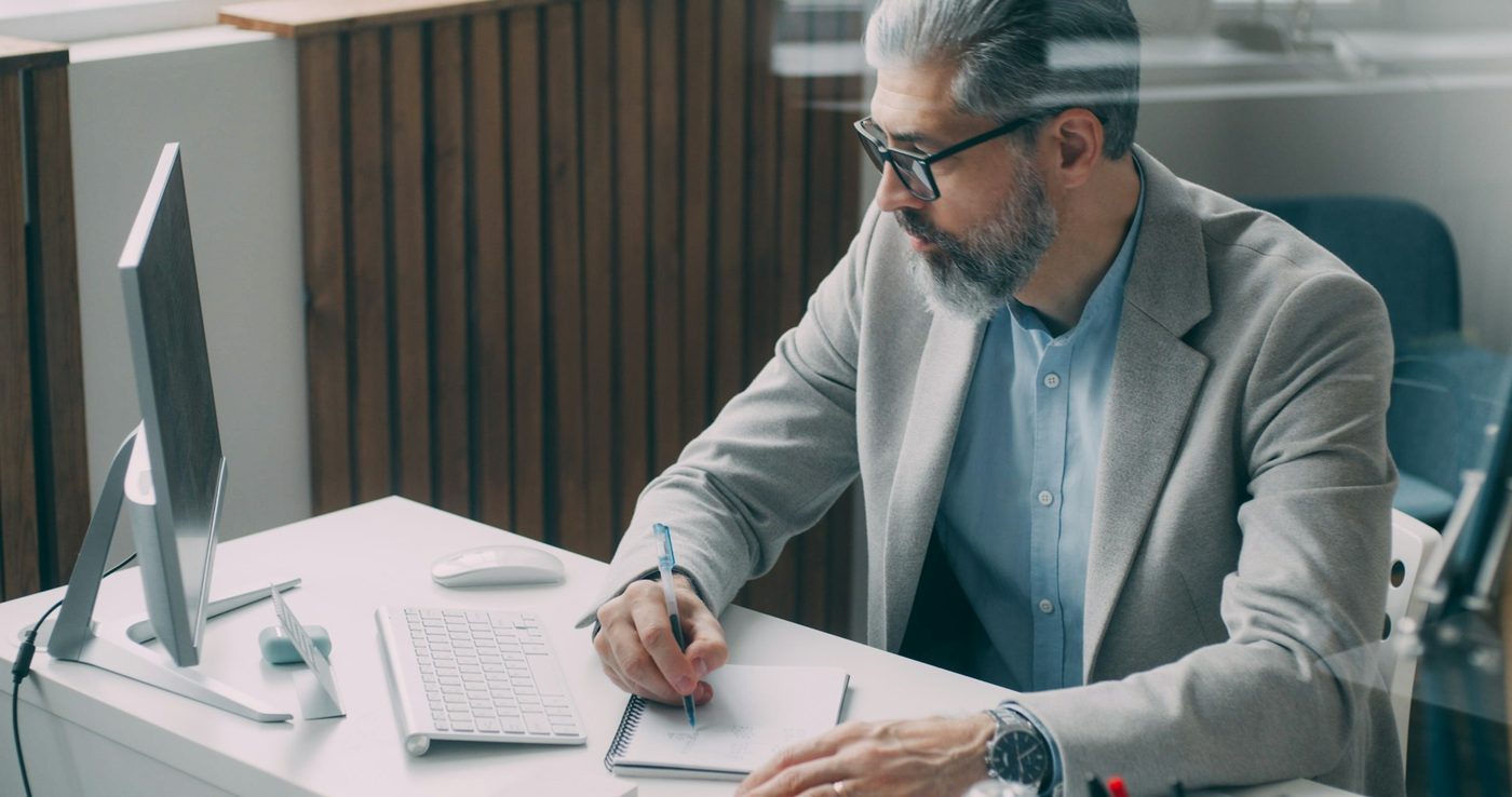 Illustrative stock photo: a man at a white desk writes on a spiral notepad with a pen. The monitor beside him is turned side-on so its screen can't be seen, and the office is lit by daylight.