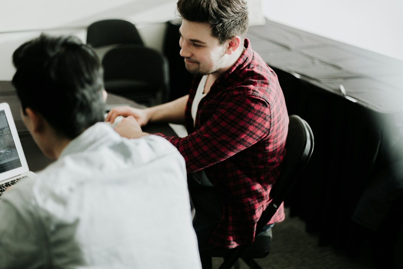 Illustrative stock photo: two colleagues in casual clothes shake hands across a table in a plain meeting room, with a laptop at the edge of the frame.