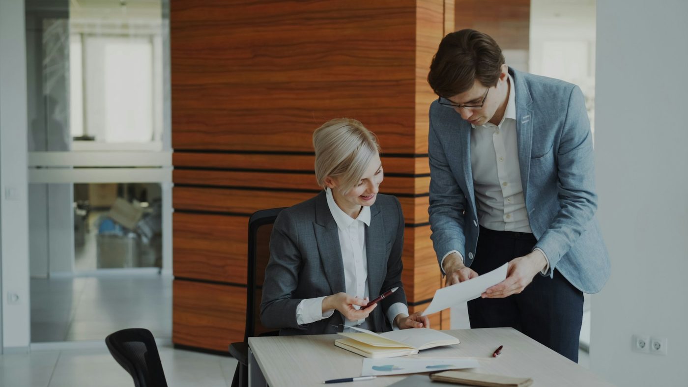 Illustrative stock photo: a woman seated at an office desk with an open notebook and papers while a colleague leans over and points at a printed page he is holding.