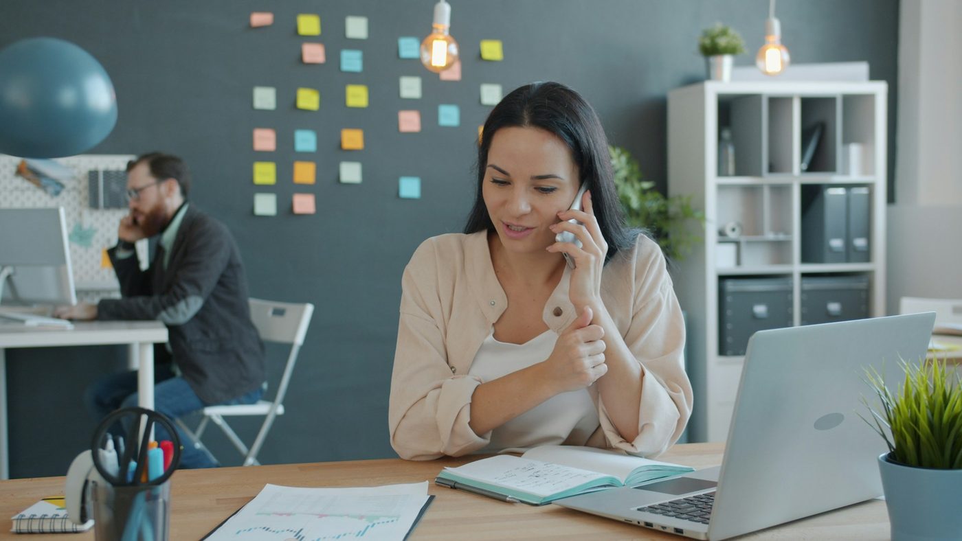 Illustrative stock photo: a woman on the phone at her desk in a small office, in front of a slate wall with a grid of coloured sticky notes, while a colleague works on a call at the next desk.
