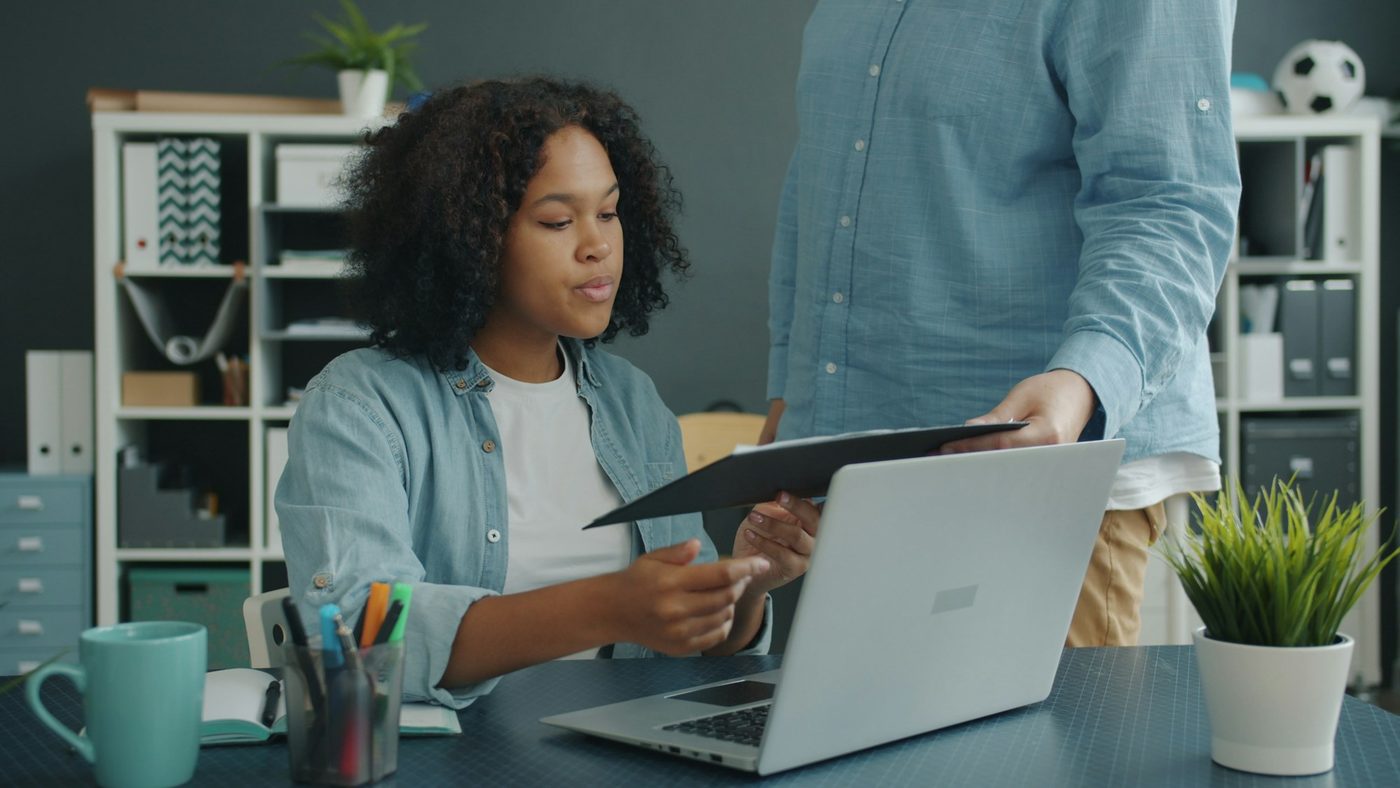 An analyst at her desk takes a document folder from a colleague standing beside her, with a laptop, a mug and pens on the desk and shelves of box files behind.