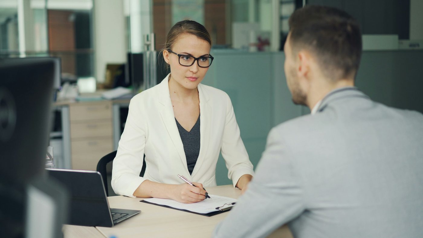 Illustrative stock photo: a woman in glasses takes notes on a clipboard during a quiet one-to-one conversation at a desk, with glass office partitions behind her and a colleague seen from behind.