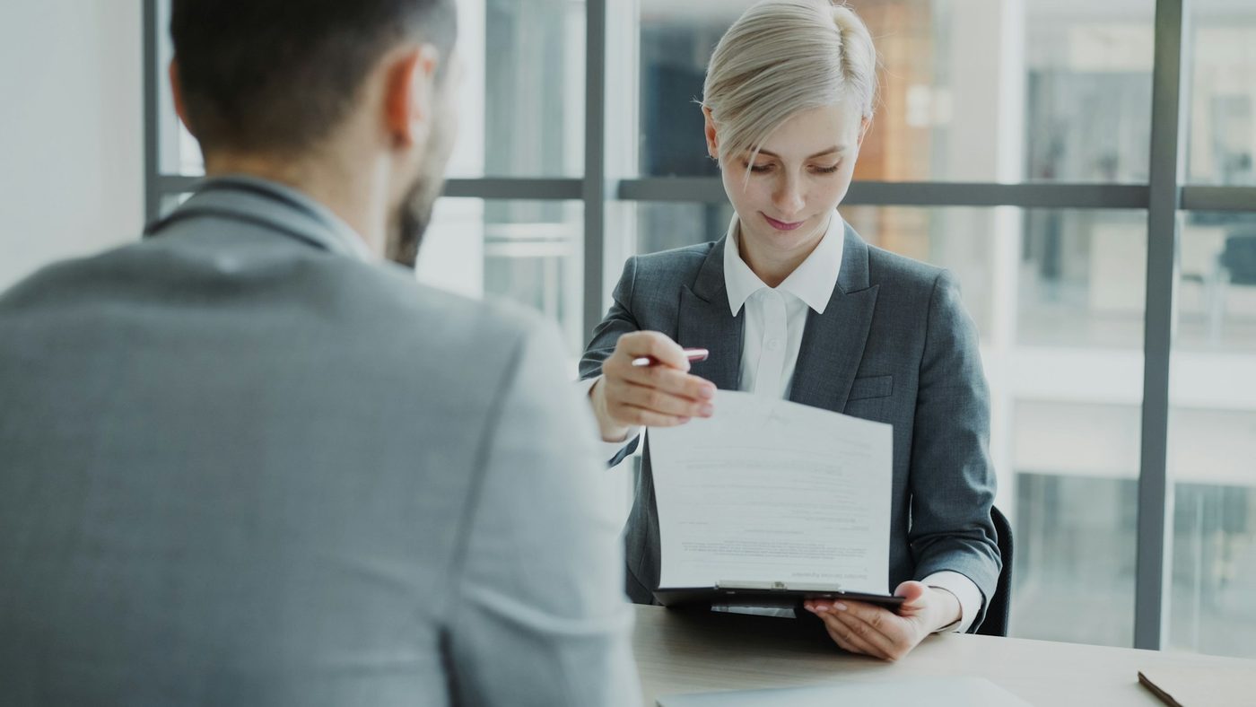A professional in a suit, pen in hand, reads a printed document on a clipboard at her desk while a colleague sits across from her.